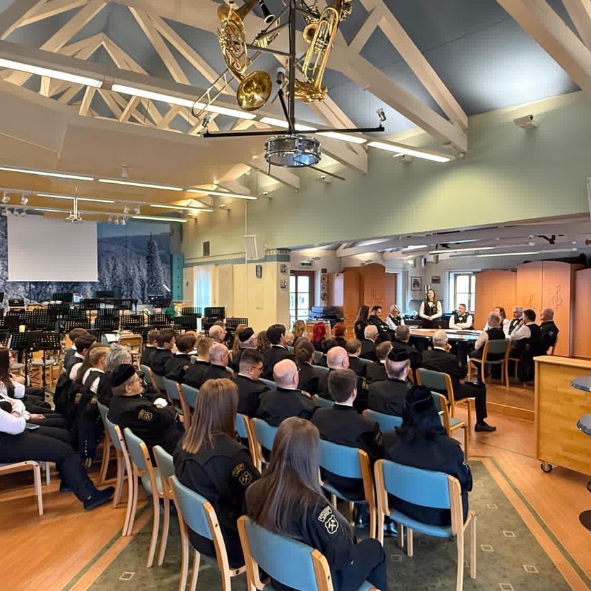 Ein Raum mit Holzboden und weißen Deckenbalken beherbergt eine Gruppe von Menschen in formeller Kleidung, die auf Stühlen sitzen und einer Vorlesung oder Präsentation lauschen. Musikinstrumente sind auf der Bühne ausgestellt.