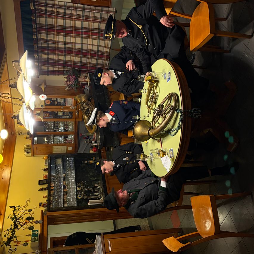 Eine Gruppe von Militäroffizieren ist um einen Tisch mit verschiedenen Messinginstrumenten versammelt, möglicherweise in einer gesellschaftlichen Umgebung. Der Raum verfügt über Holzstühle, eine Deckenleuchte und eine gelbe Wand.