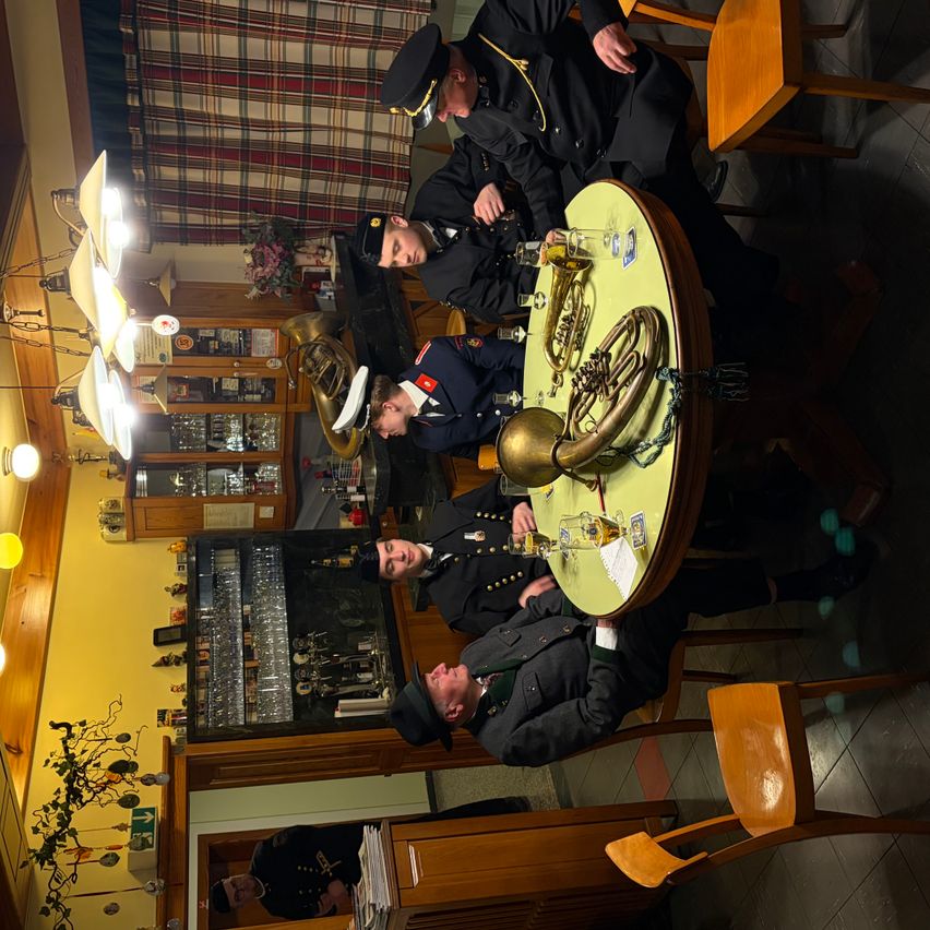 Eine Gruppe von Militäroffizieren versammelt sich um einen Tisch, vermutlich in einer Bar, mit einem großen Messinginstrument darauf. Sie scheinen ein Gespräch zu führen und zu trinken. Der Raum hat gelbe Wände, Holzstühle und einen Kronleuchter.