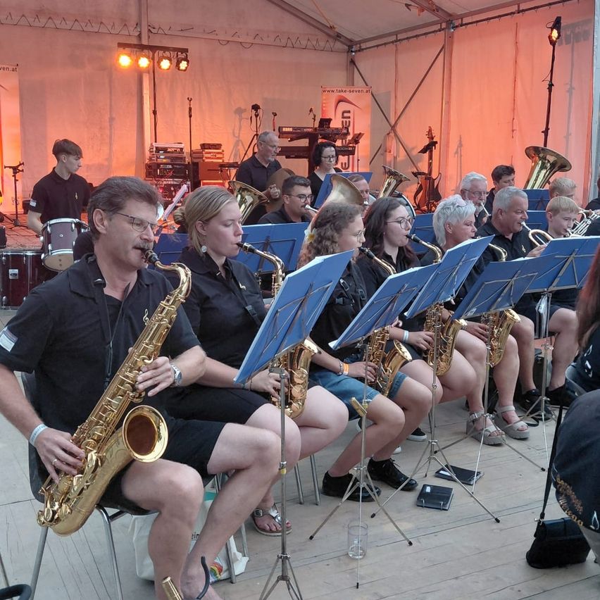 Eine Gruppe von Musikern spielt auf der Bühne mit verschiedenen Instrumenten, darunter Saxophone und Trompeten. Die Musiker sitzen und stehen, einige halten ihre Instrumente. Die Bühne ist gut beleuchtet, und es gibt Notenständer und Ausrüstung im Hintergrund.