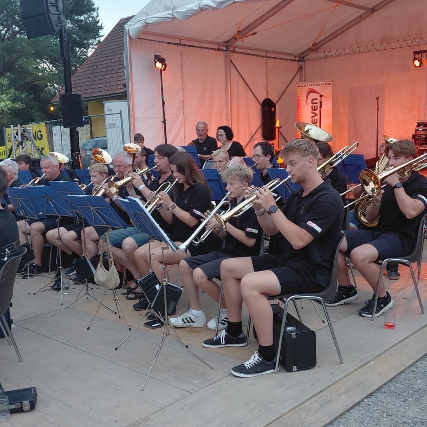 Ein Blasorchester spielt unter einem Zelt, wobei die Musiker in Stühlen sitzen und Trompeten und Posaunen halten. Im Hintergrund ist ein Banner mit dem Wort 'EVEN' zu sehen.