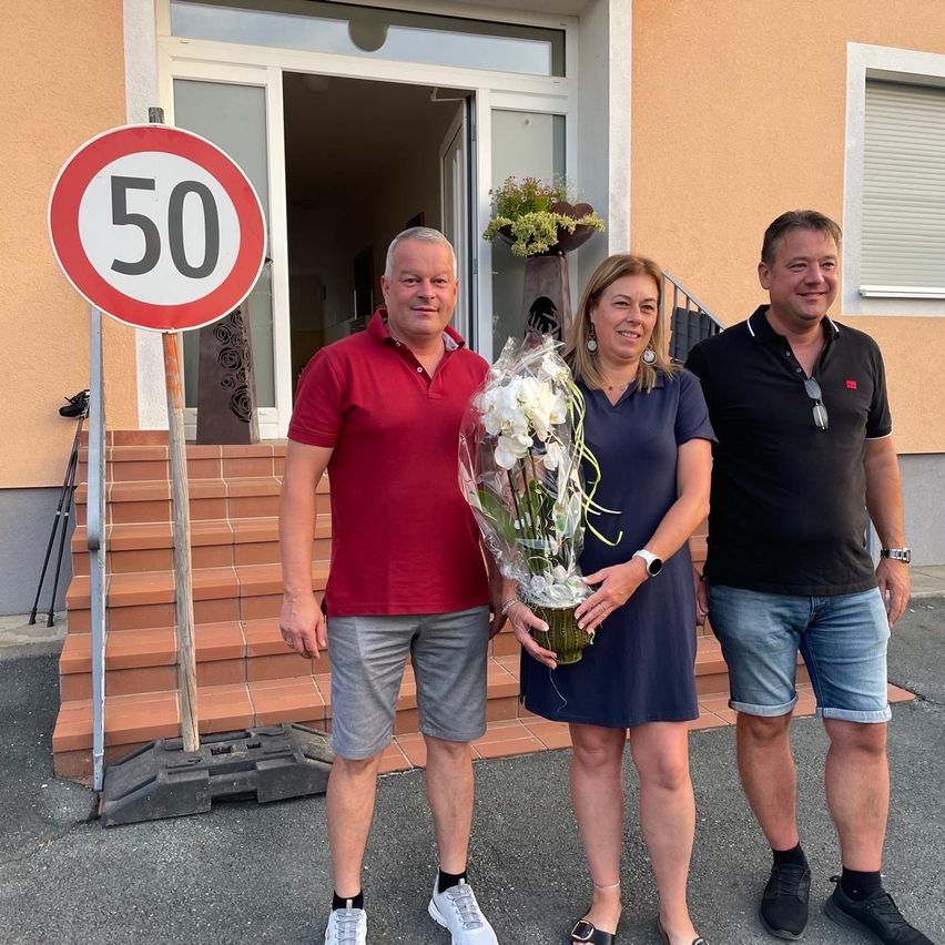 Three people stand in front of a building with a 50 km/h speed limit sign. A man in red holds a bouquet of flowers, while a woman in blue smiles. A man in black stands beside them.