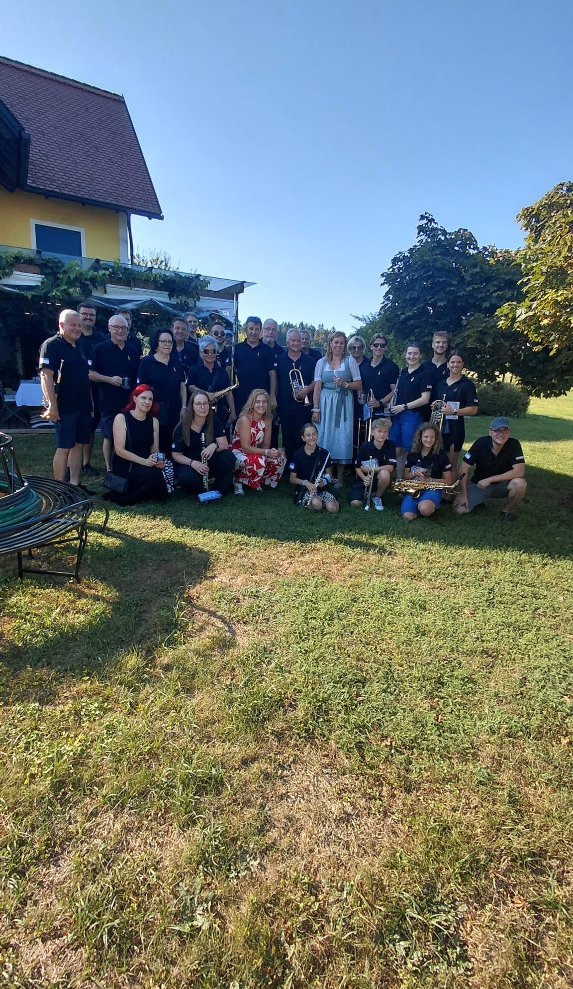 A group of musicians in black shirts pose for a photo in a grassy field with a house in the background.