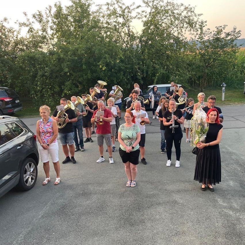 A group of musicians in a parking lot, holding brass instruments, with a woman in a black dress holding flowers in the foreground.
