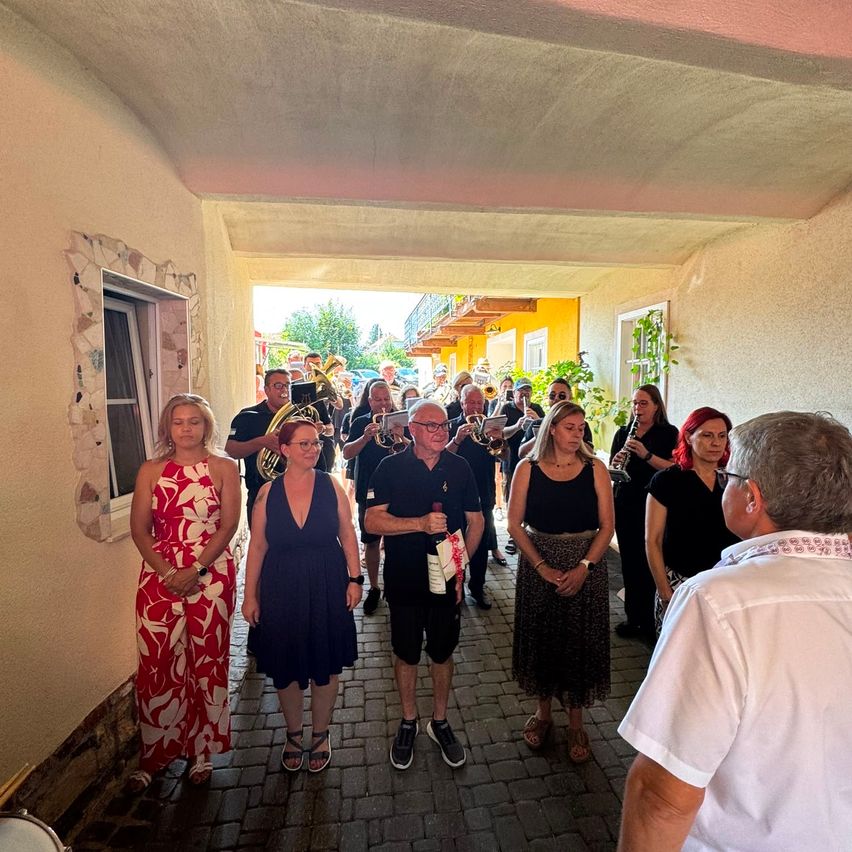 Eine Gruppe von Menschen, darunter Frauen in Kleidern und Männer in Freizeitkleidung, steht in einem Korridor, während eine Band hinter ihnen spielt. Die Bandmitglieder halten Blechblasinstrumente.