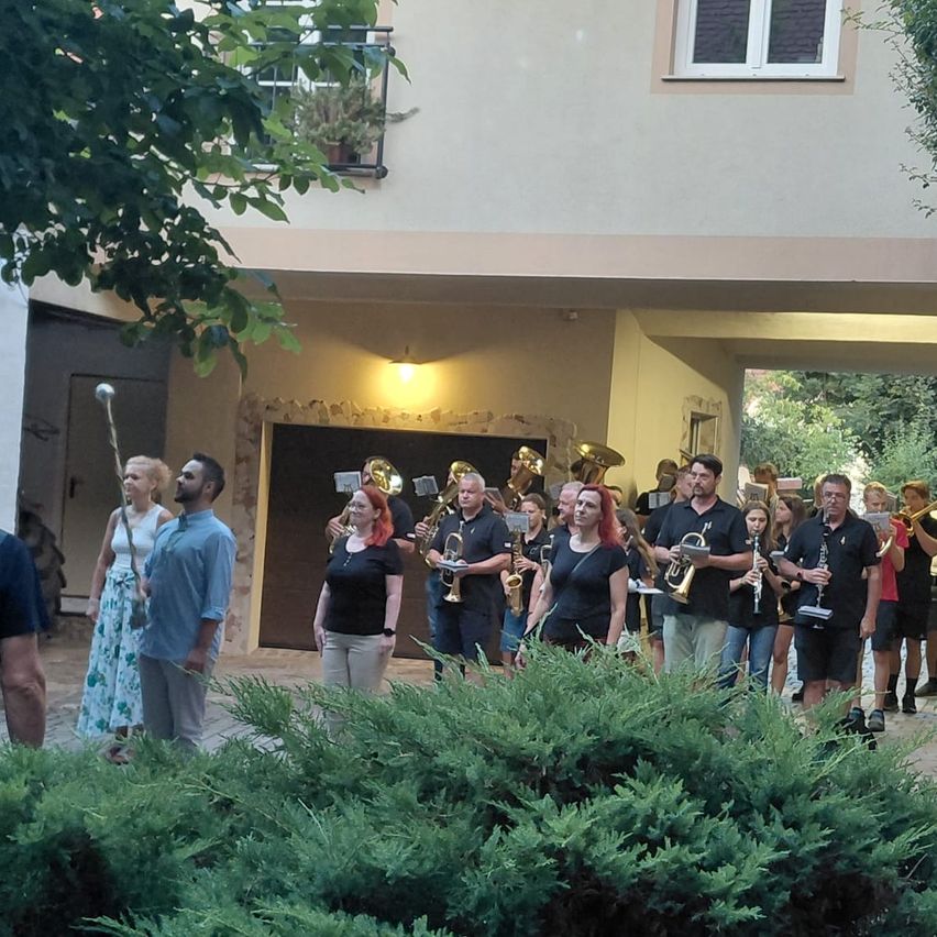 Eine Gruppe von Musikern, darunter Trompeten und Posaunen, steht vor einem Gebäude mit Balkon und Fenster.