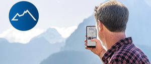 Ein Mann und ein Junge schauen auf ein Smartphone im Freien. Der Junge hat seinen Arm um die Schulter des Mannes gelegt. Hinter ihnen ist eine Bergkette.