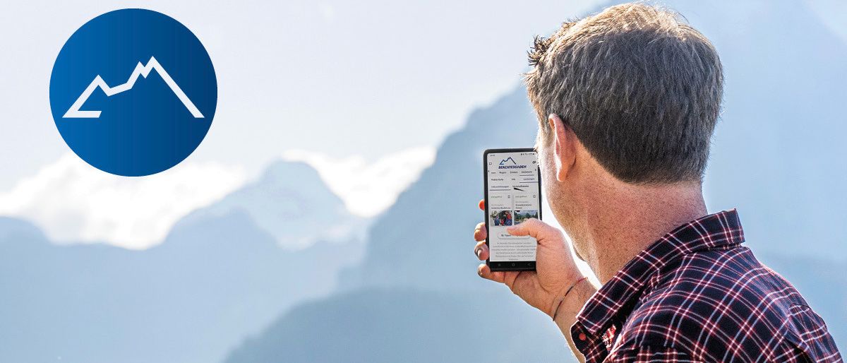 Ein Mann und ein Junge schauen auf ein Smartphone im Freien. Der Junge hat seinen Arm um die Schulter des Mannes gelegt. Hinter ihnen ist eine Bergkette.