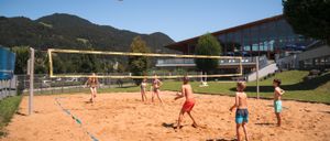Mehrere Kinder spielen Beachvolleyball auf einem Sandplatz. Der Ball ist in der Luft, und die Spieler stehen um das Netz herum. Dahinter befindet sich ein Gebäude mit Terrasse und Bergen im Hintergrund.