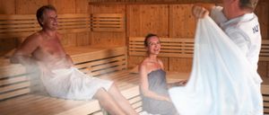 Ein älterer Mann und eine Frau sitzen lächelnd in einer Holzsauna. Der Mann trägt ein weißes Handtuch, die Frau ein graues. Eine Person hinter ihnen hält ein Handtuch.