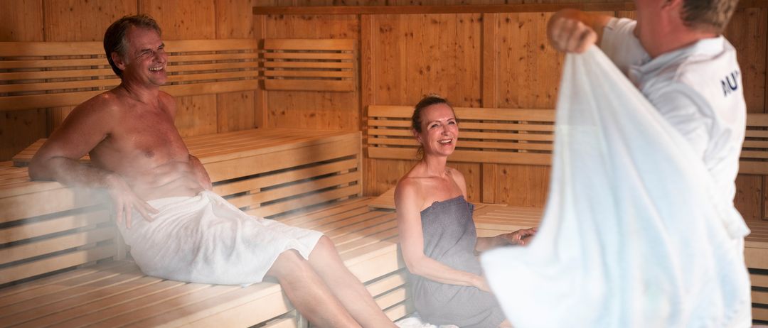 Ein älterer Mann und eine Frau sitzen lächelnd in einer Holzsauna. Der Mann trägt ein weißes Handtuch, die Frau ein graues. Eine Person hinter ihnen hält ein Handtuch.