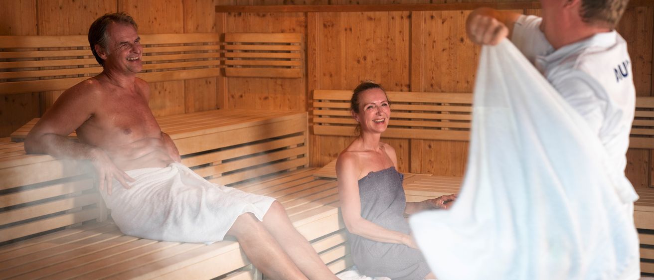 Ein älterer Mann und eine Frau sitzen lächelnd in einer Holzsauna. Der Mann trägt ein weißes Handtuch, die Frau ein graues. Eine Person hinter ihnen hält ein Handtuch.