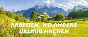 Eine saftige grüne Wiese mit bunten Blumen und einer Bergkette im Hintergrund. Der Textüberlagerung lautet 'Arbeiten, wo andere Urlaub machen'.