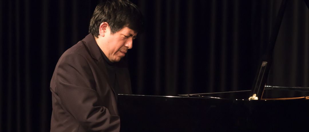 Ein Mann spielt geschlossenäugig auf der Bühne Klavier. Er trägt einen dunklen Anzug. Hinter ihm befindet sich ein schwarzer Vorhang.