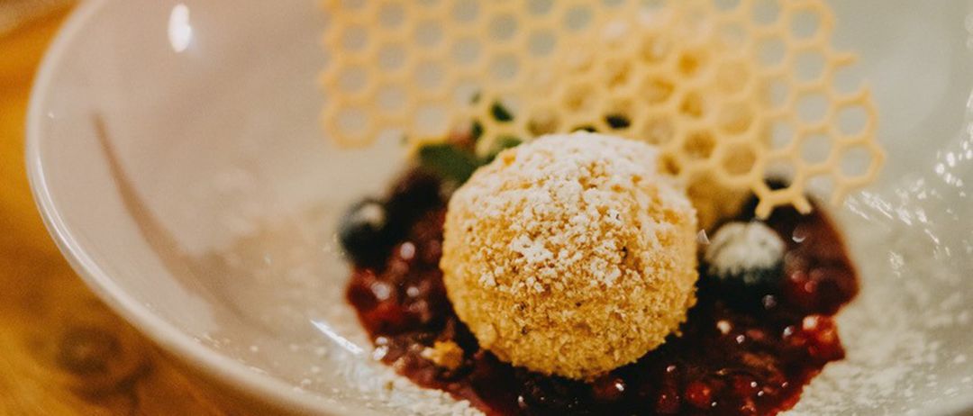 Ein Dessert in einer weißen Schüssel mit einer goldbraunen Kugel, bedeckt mit Puderzucker, umgeben von einer dunklen Beerensauce und einer honigwabenförmigen Garnitur.