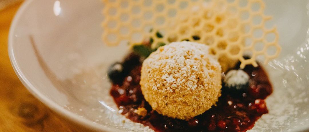 Ein Teller mit Dessert zeigt eine goldene Kugel, bestreut mit Zucker, begleitet von einer roten Beerensauce und garniert mit Blaubeeren und einem honigwabenförmigen Element.