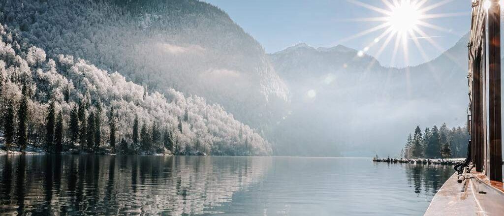 Ein ruhiger See umgeben von verschneiten Bergen unter einer strahlenden Sonne. Das Wasser reflektiert die Sonnenstrahlen und der Himmel ist klar.