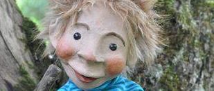 Eine kleine Puppe mit blonden Haaren, die ein blau gestreiftes Shirt trägt, sitzt auf einem Ast und hält einen Stock in der Hand.