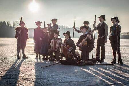 Eine Gruppe von Musikern in traditioneller Kleidung steht auf einem gefrorenen See und spielt Instrumente wie Flöten, Gitarren und Akkordeons unter einem strahlenden Himmel.