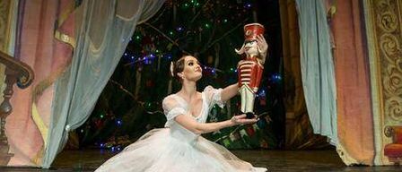 Eine Ballerina in einem weißen Kleid kniet auf der Bühne und hält eine Nussknackerfigur. Hinter ihr steht ein Weihnachtsbaum und prunkvolle Vorhänge.
