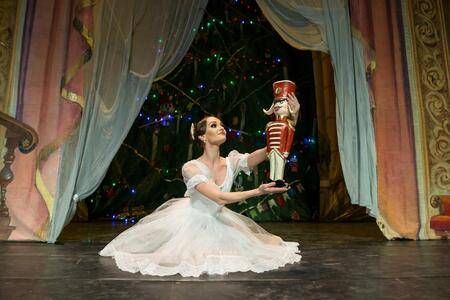 Eine Ballerina in einem weißen Kleid kniet auf der Bühne und hält eine Nussknackerfigur. Hinter ihr steht ein Weihnachtsbaum und prunkvolle Vorhänge.