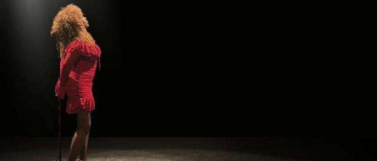 Eine Frau in einem roten Kleid und hohen Absätzen steht auf einer Bühne, beleuchtet von Scheinwerfern. Sie scheint zu singen.