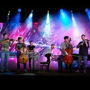 Eine Gruppe von Musikern tritt auf einer Bühne auf, mit leuchtenden Lichtern im Hintergrund, und spielt Cello, Geige und Klavier.