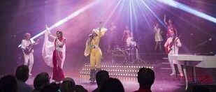 Eine lebhafte Bühnenshow mit Musikern in farbenfrohen Kostümen unter hellen Scheinwerfern, die ein begeistertes Publikum vor der Bühne unterhalten.