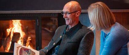 Ein älterer Mann mit Brille sitzt mit einer Frau in der Nähe eines Kamins, beide schauen in ein Buch, das der Mann in der Hand hält. Das Feuer brennt im Hintergrund hell.