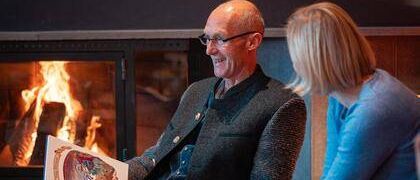 Ein älterer Mann mit Brille liest einer neben ihm sitzenden Frau ein Buch vor, vor einem Kamin.