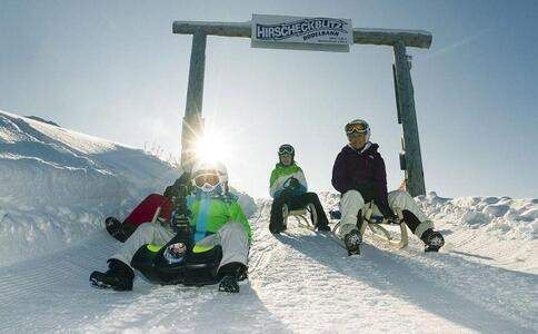 Drei Personen sitzen auf einem verschneiten Hang in Schlitten mit einem Holzschild, auf dem Hirscheckblitz steht. Sie tragen Schutzbrillen und Helme und scheinen für ein Foto zu posieren.