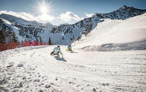 Zwei Personen rutschen bei strahlendem Sonnenlicht einen schneebedeckten Berg hinunter. Bäume und Berge sind im Hintergrund zu sehen.