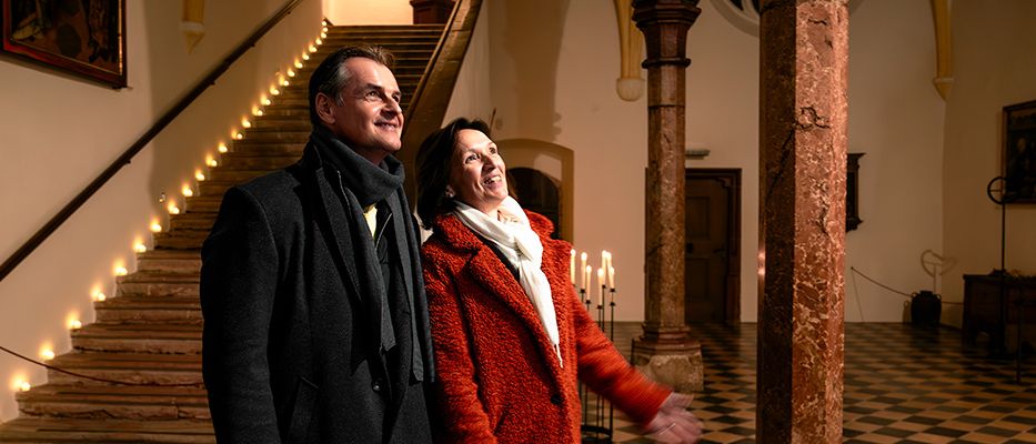 Ein Mann und eine Frau stehen lächelnd neben einer Treppe mit brennenden Kerzen in einem historischen Gebäude mit Marmorsäulen und einem gescheckten Boden.