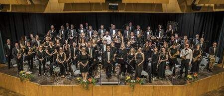 Ein großes Orchester posiert für ein Gruppenfoto auf einer mit Blumen geschmückten Bühne, mit leeren Stühlen darunter.