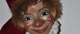 Ein Nahaufnahme einer bemalten Holzpuppe mit roter Kapuze und weißer Spitzenkrawatte. Die Puppe hat ein bemaltes Gesicht mit roter Nase und roten Wangen.