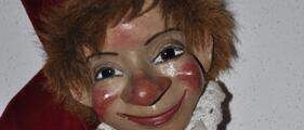A doll dressed in red, featuring a red hat, white ruffled collar, and painted-on clown makeup. The doll has brown hair, red cheeks, and a big red nose.