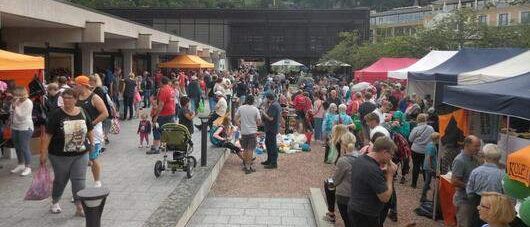Ein überfüllter Außenmarkt mit Menschen, die herumlaufen, einige mit Kinderwagen, vor verschiedenen Zelten und einem Gebäude. Bäume und Berge im Hintergrund.