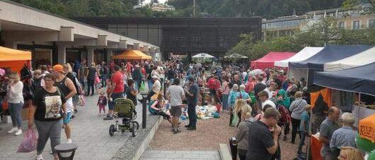 Ein belebter Freiluftmarkt mit vielen Menschen, die herumlaufen, einige mit Kinderwagen und Taschen. Zelte mit bunten Abdeckungen sind aufgebaut, und ein Gebäude ist im Hintergrund sichtbar.