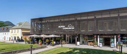 Der Eingang des Alpen Congress Hotels mit einer modernen Fassade, einem Rasen und einem Außenbereich mit Tischen, Sonnenschirmen und Pflanzen.