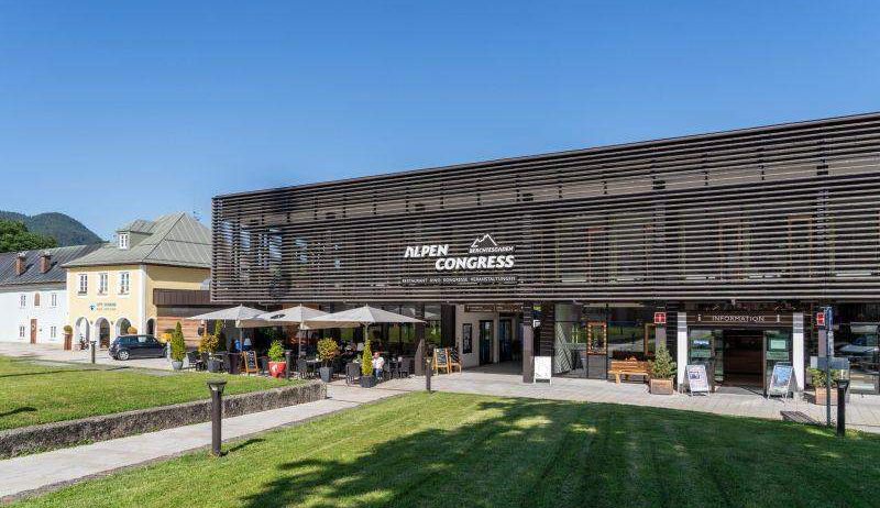 Der Eingang des Alpen Congress Hotels mit einer modernen Fassade, einem Rasen und einem Außenbereich mit Tischen, Sonnenschirmen und Pflanzen.