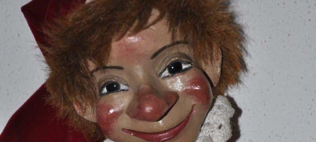 Ein Nahaufnahme des Gesichts einer Puppe, die einen roten Hut und einen roten und weißen Schal trägt, mit einer bemalten roten Nase und roten Wangen.