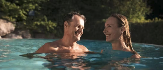 Ein Mann und eine Frau schwimmen in einem Pool, lächeln und schauen sich an. Bäume und Felsen sind im Hintergrund.