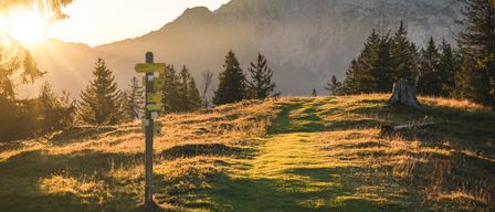 Ein Wanderwegschild in einem Grasfeld mit einem Bergrücken im Hintergrund während des Sonnenuntergangs.