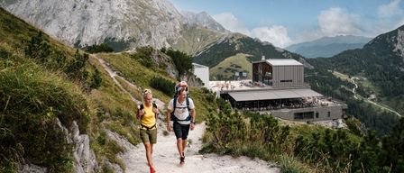 Zwei Wanderer gehen auf einem Bergpfad, mit einem Gebäude und Bergen im Hintergrund.