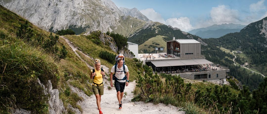 Zwei Wanderer gehen auf einem Bergpfad, mit einem Gebäude und Bergen im Hintergrund.