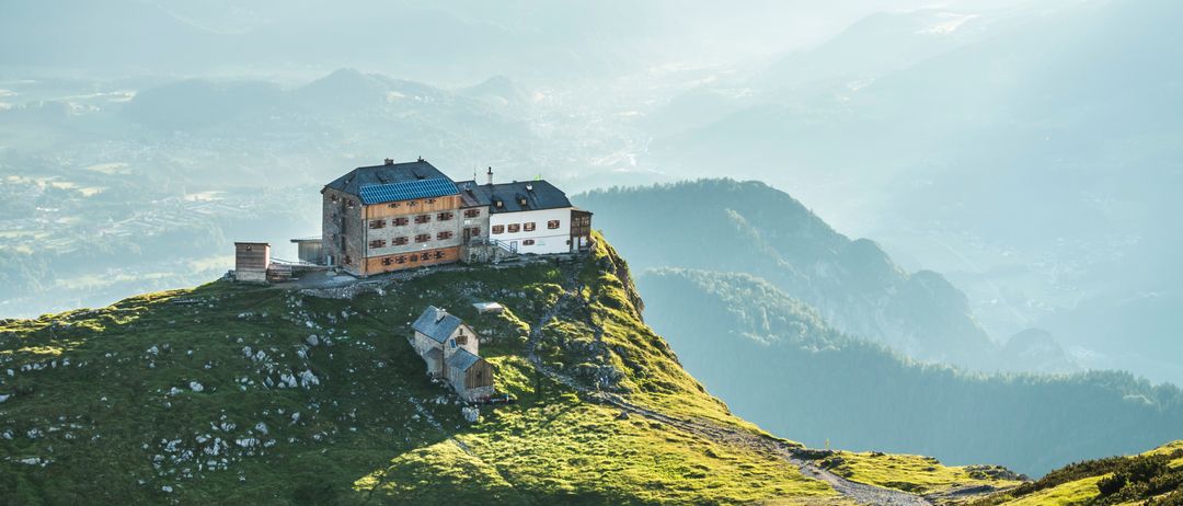 Eine Berghütte mit blauem Dach und Solarpanels steht auf einem grasbewachsenen Hügel, umgeben von üppigen grünen Bergen.