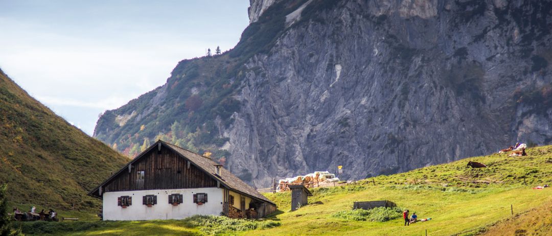 A traditional wooden house stands on a grassy hillside with a steep rocky cliff in the background. Two people are walking on the path in the foreground.