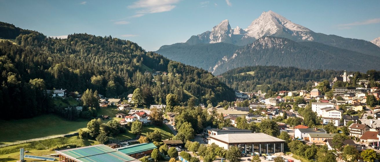 Luftaufnahme eines Bergdorfes mit mehreren Gebäuden und einem großen Gebäude mit grünem Dach. Berge im Hintergrund und blauer Himmel.