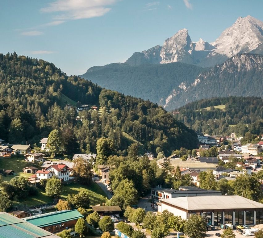 Luftaufnahme eines Bergdorfes mit mehreren Gebäuden und einem großen Gebäude mit grünem Dach. Berge im Hintergrund und blauer Himmel.