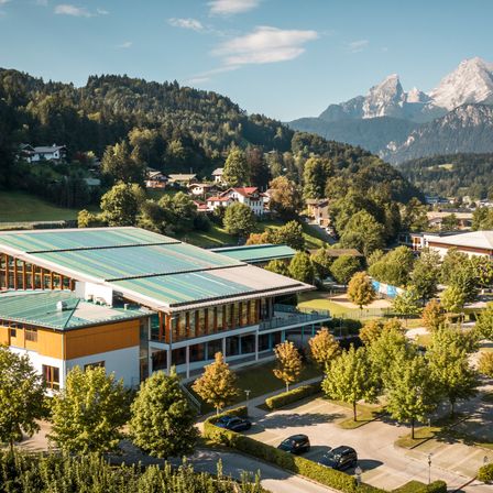 Ein Luftbild eines modernen Gebäudes mit grünen Dächern und Solarmodulen. Das Gebäude ist von Bäumen und Büschen umgeben. In der Ferne befinden sich Häuser und Berge.